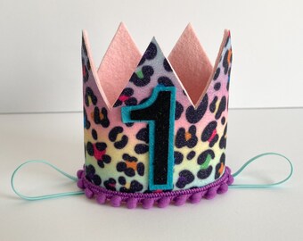 May include: A pink and rainbow leopard print felt crown with a black felt number one and a purple pom-pom trim. The crown has a light blue elastic band.