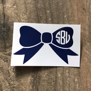 May include: A navy blue bow-shaped sticker with a white background. The bow features a monogram with the letters "SBL" in a circular design. The sticker is on a wooden surface.