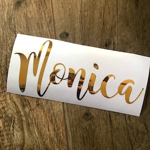 Op de afbeelding: Een gouden metallic sticker met de naam "Monica" in een sierlijk handschrift. De sticker bevindt zich op een witte rechthoekige achtergrond, tegen een houten oppervlak. De gouden letters reflecteren de omgeving.