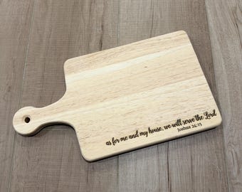 Puede incluir: Una tabla de cortar de madera clara con un mango que tiene un orificio para colgar. La tabla está grabada con el texto: as for me and my house, we will serve the Lord Joshua 24:15 en una letra cursiva oscura en la parte inferior derecha.
