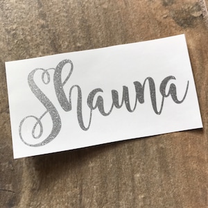 Pode incluir: Um autocolante retangular branco com o nome "Shauna" em escrita prateada brilhante. O autocolante está numa superfície castanha texturizada.