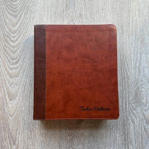 May include: A rectangular brown binder with a darker brown vertical strip along the left edge. The cover features a smooth, textured finish and the name Tucker Hallman is embossed in a dark, elegant script font in the bottom right corner.