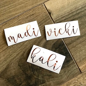 May include: Three rectangular white adhesive labels featuring custom names in a metallic rose gold script font. The labels display the names madi, vicki, and kali in a flowing, cursive typeface with a reflective finish on a smooth white background.