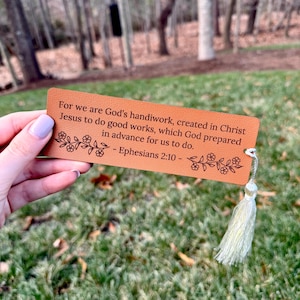 Könnte beinhalten: Ein rechteckiger, hellbrauner Leder-Lesezeichen mit dem Text "For we are God's handiwork, created in Christ Jesus to do good works, which God prepared in advance for us to do. - Epheser 2:10 -" und floralen Designs. Eine weiße Quaste hängt oben.
