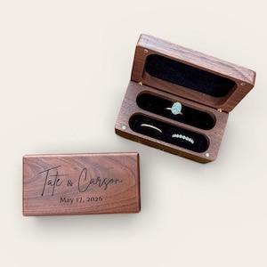 Peut inclure: Un écrin à bagues en bois, ouvert pour révéler trois fentes doublées de velours noir. La boîte est gravée des noms "Tate & Carson" et de la date "17 mai 2026". La boîte contient trois bagues. Le bois est foncé.