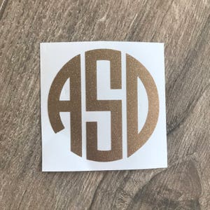 May include: A gold glitter monogram sticker with the letters ASD in a circular design. The sticker is on a white background and is placed on a wooden surface.