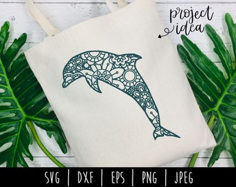 May include: A tote bag with a teal mandala dolphin design and the text "project idea" on the top right corner. The bag is on a white wooden surface with green leaves.