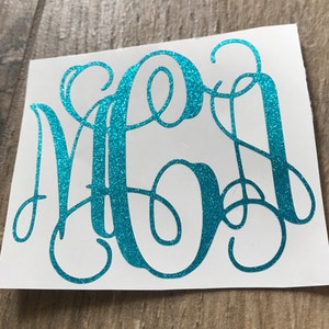 May include: A teal glitter monogram sticker featuring the letters M, A, and O in a cursive font. The sticker is on a white background, with a wooden surface visible in the background.