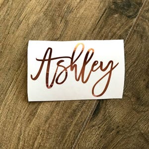 May include: A white sticker with the name "Ashley" in a copper-colored, cursive font. The sticker is rectangular and sits on a brown wooden surface.