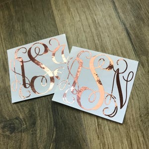 May include: Two square decals with rose gold monogram initials. The letters are stylized with elegant swirls and flourishes. The decals are on a light blue background.