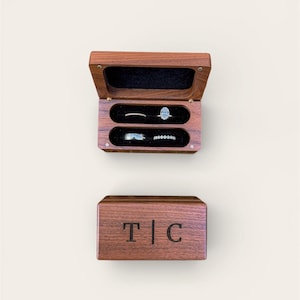 May include: A wooden ring box, open to reveal two compartments with black velvet lining, each holding a ring. The top of the box is open, displaying an oval diamond ring and a thin band. The closed box below has the initials "T | C".