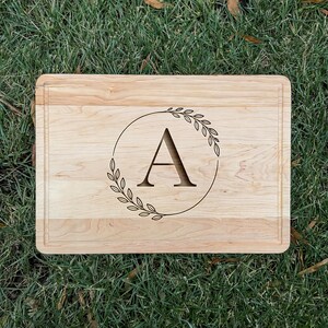 Könnte beinhalten: Ein rechteckiges Holzschneidebrett mit einer Saftrille. Das Brett zeigt einen großen Buchstaben "A" in einem kreisförmigen Kranzdesign. Das Holz hat eine helle, natürliche Farbe und das Design ist in das Holz geätzt.