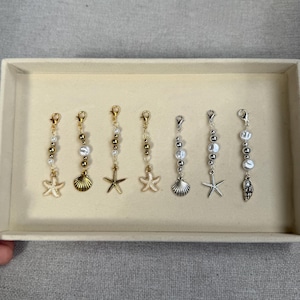 May include: Seven beach-themed charms with lobster claw clasps. The charms feature gold and silver tones, with starfish, seashells, and pearl accents. The charms are displayed in a beige box.