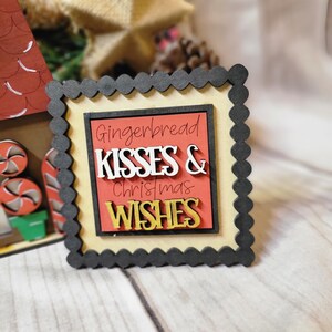 May include: A black framed sign with a scalloped edge. The sign has a cream background with a red rectangle in the center. The text "Gingerbread Kisses & Christmas Wishes" is written in white and gold letters.