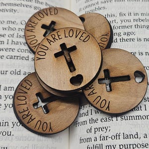 May include: A set of six wooden round ornaments with a cross and heart cutouts. Each ornament has the text "You Are Loved" written around the edge.