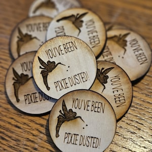 May include: A collection of round wooden tokens with a fairy illustration and the text "YOU'VE BEEN PIXIE DUSTED!". The tokens are light brown with a darker, burnt-in design. The fairy is depicted with outstretched wings.