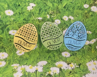 May include: Three wooden Easter egg ornaments with the name "Oliver" engraved on each. The eggs are decorated with different patterns: a woven pattern, a dotted pattern, and a floral pattern. The eggs are light brown, light green, and light blue.