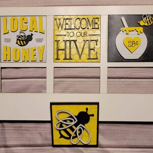 May include: White wooden sign with four square panels. The top left panel says "LOCAL HONEY" with a bee and honeycomb design. The top middle panel says "WELCOME TO OUR HIVE" with a honeycomb design. The top right panel has a black and yellow bee and a black honey pot with a white heart that says "25c". The bottom panel has a bee with a honeycomb design.
