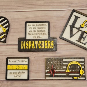 May include: A collection of wooden decorative pieces with a law enforcement theme. Includes a heart, a sign with the word "DISPATCHERS", a square with the word "LOVE" and a 911 symbol, a square with the text "In our Family, no one fights alone", and a flag design.