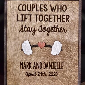 May include: Wooden sign with the text "COUPLES WHO LIFT TOGETHER Stay Together". Features a barbell with a heart in the middle. Below the barbell is the text "MARK AND DANIELLE April 24th, 2023".
