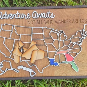 Puede incluir: Mapa de madera de los Estados Unidos con un marco marr&oacute;n oscuro. El mapa presenta contornos blancos de los estados y acentos coloridos. El texto "Adventure Awaits" y "NOT ALL WHO WANDER ARE LOST" se muestra en blanco.