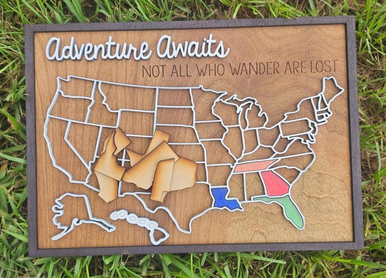 Puede incluir: Mapa de madera de los Estados Unidos con un marco marr&oacute;n oscuro. El mapa presenta contornos blancos de los estados y acentos coloridos. El texto "Adventure Awaits" y "NOT ALL WHO WANDER ARE LOST" se muestra en blanco.