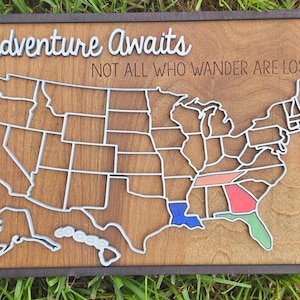 May include: Wooden map of the United States with state outlines in white, set against a brown background. The words "Adventure Awaits" and "NOT ALL WHO WANDER ARE LOST" are displayed. Some states are colored in blue, red, green, and orange.