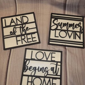 May include: Three white wooden signs with black lettering. The signs read "Land of the Free", "Summer Lovin'" and "Love Begins at Home".