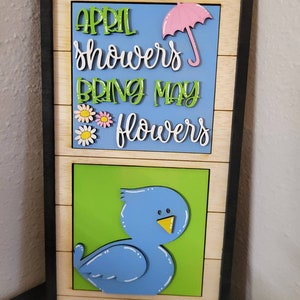 May include: A wooden sign with the saying "April showers bring May flowers" in colorful letters. The sign also features a pink umbrella and a blue bird.