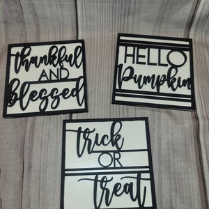 May include: Three square wooden signs with black borders and white backgrounds. The signs feature black cursive text that reads "thankful AND blessed", "HELLO Pumpkin", and "trick OR treat". These signs are perfect for fall and Halloween decor.