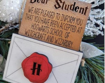 Puede incluir: Un adorno de madera con forma de sobre con un sello de lacre rojo que dice "H". El sobre está unido a una tarjeta de madera que dice "Estimado estudiante, Nos complace informarle que ha sido aceptado en el Colegio Hogwarts de Magia y Hechicería."