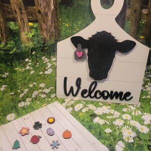 May include: Wooden cutout of a cow with a pink heart tag, hanging on a white wooden sign that says "Welcome".  The sign is in front of a green grassy area with white flowers.  There are 10 small wooden ornaments with various designs on a white wooden surface.