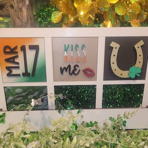 May include: A white wooden frame with a green, orange, and white background. The frame has six squares with different images. The top left square says "MAR 17". The top middle square says "KISS ME". The top right square has a gold horseshoe with a green shamrock. The bottom left square has a green shamrock. The bottom middle square has green glitter. The bottom right square has green glitter.