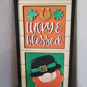 May include: A wooden sign with a black frame and a woodgrain background. The sign has a layered design with a top section featuring a gold horseshoe, green shamrocks, and the text "lucky & blessed" in white lettering on an orange background. The bottom section features a cartoon leprechaun with a green hat, orange beard, and a brown belt.