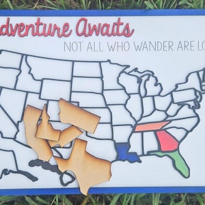 Puede incluir: Mapa de madera de los Estados Unidos con piezas de estado faltantes. Las palabras "Adventure Awaits" y "NOT ALL WHO WANDER ARE LOST" est&aacute;n escritas en el fondo blanco. El marco es azul.