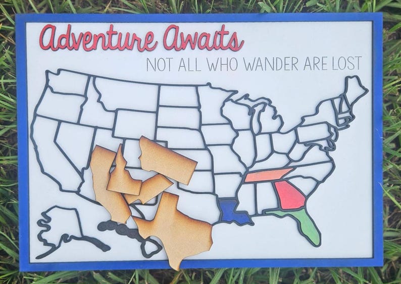 Puede incluir: Mapa de madera de los Estados Unidos con piezas de estado faltantes. Las palabras "Adventure Awaits" y "NOT ALL WHO WANDER ARE LOST" est&aacute;n escritas en el fondo blanco. El marco es azul.