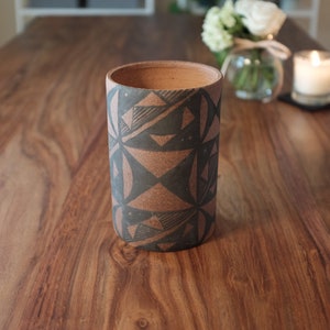 May include: A brown ceramic vase with a geometric black and white pattern. The vase is sitting on a wooden table.