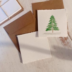 K&ouml;nnte beinhalten: Drei leere Karten mit braunen Umschl&auml;gen. Eine Karte hat ein Aquarellbild eines gr&uuml;nen Weihnachtsbaums.