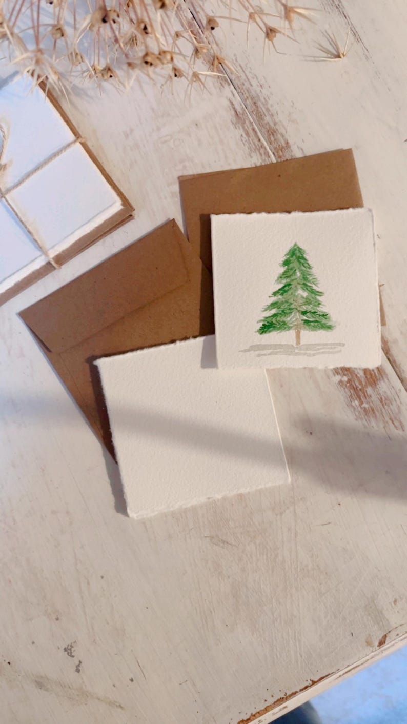 K&ouml;nnte beinhalten: Drei leere Karten mit braunen Umschl&auml;gen. Eine Karte hat ein Aquarellbild eines gr&uuml;nen Weihnachtsbaums.
