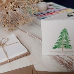 K&ouml;nnte beinhalten: Eine wei&szlig;e Karte mit einem Aquarellbild eines gr&uuml;nen Weihnachtsbaums auf einem wei&szlig;en Holztisch. Im Hintergrund befinden sich ein Stapel wei&szlig;en Papiers und ein brauner Umschlag.