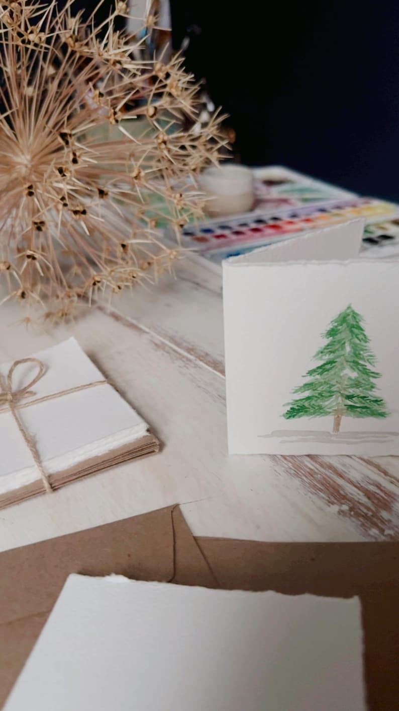 K&ouml;nnte beinhalten: Eine wei&szlig;e Karte mit einem Aquarellbild eines gr&uuml;nen Weihnachtsbaums auf einem wei&szlig;en Holztisch. Im Hintergrund befinden sich ein Stapel wei&szlig;en Papiers und ein brauner Umschlag.