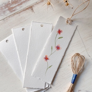 May include: Four white paper bookmarks with a delicate floral design painted in shades of pink and orange. The bookmarks are stacked on a white wooden surface with a piece of twine and a blue tag.