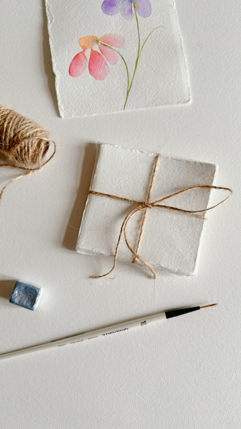 K&ouml;nnte beinhalten: Ein Stapel wei&szlig;er Papierkarten, die mit Schnur zusammengebunden sind, ein Aquarellbild von Blumen, ein Pinsel und ein kleiner blauer Farbkubus.