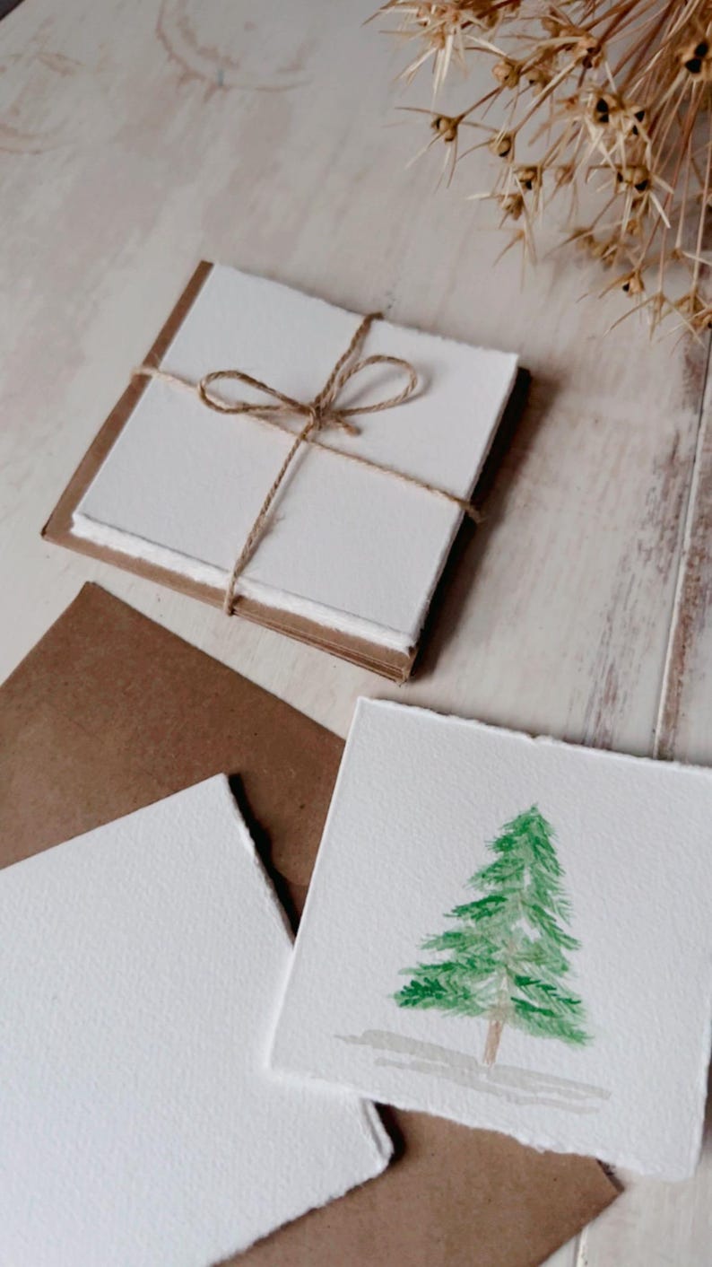 K&ouml;nnte beinhalten: Ein Satz leerer Karten mit einem braunen Papierumschlag. Die Karten sind mit Schnur zusammengebunden und haben ein rustikales, handgemachtes Aussehen. Eine Karte zeigt ein Aquarellbild eines gr&uuml;nen Weihnachtsbaums.