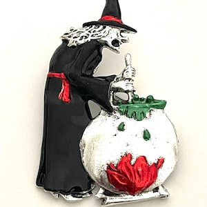 May include: A Halloween-themed brooch featuring a witch stirring a cauldron. The witch is dressed in a black robe with a red sash and a pointed hat. The cauldron is silver with green and red accents. The brooch is made of metal.