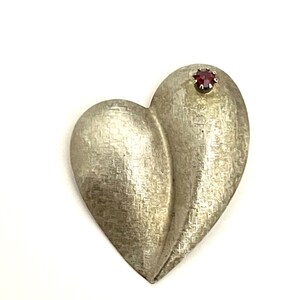 May include: A silver heart-shaped brooch with a textured surface and a single red rhinestone in the center.