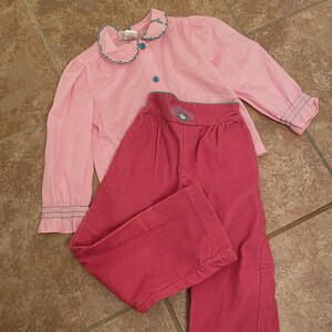Op de afbeelding: Een lichtroze shirt met lange mouwen, een geschulpte kraag met blauwe rand en blauwe knopen, gecombineerd met een donkerroze corduroy broek met hoge taille, een blauw geborduurd bloemdetail en wijde pijpen.