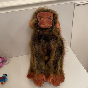 May include: A plush monkey toy featuring long, shaggy brown and black faux fur, a smooth brown face with dark eyes, and large brown hands and feet. The stuffed animal has a small pink tag attached to the side of its head and a soft, upright posture.