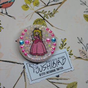 Puede incluir: Un amuleto de resina blanca con un fondo de purpurina rosa y blanca. El amuleto presenta una imagen de dibujos animados de la princesa Peach de la serie de videojuegos Super Mario Bros. El amuleto está rodeado de acentos de purpurina rosa y azul. El amuleto está hecho por Jushibird, una empresa de arte digital y resina.