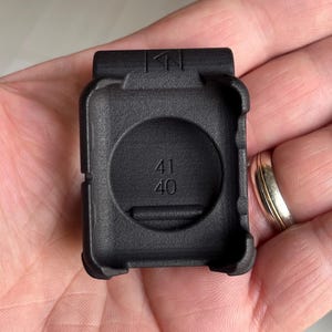 May include: A black protective case for a smartwatch, featuring a textured matte finish and a rectangular frame. The interior of the case is embossed with the numbers 41 and 40, and the top edge displays a small geometric logo symbol.